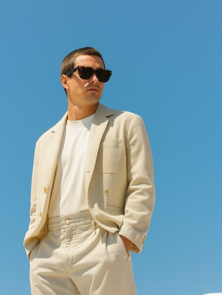 Uomo adulto in giacca e pantaloni beige chiaro con T-shirt bianca e occhiali da sole, in piedi davanti a un cielo terso e azzurro.