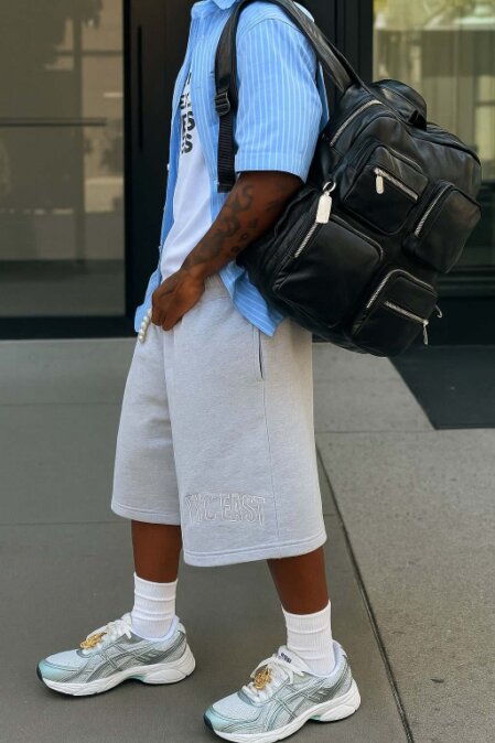 Persona in abbigliamento casual con camicia azzurra a righe sopra una felpa bianca, pantaloncini grigi e scarpe sportive, con grande zaino nero a più tasche appoggiato sulla spalla, ritratta in piedi su sfondo urbano.