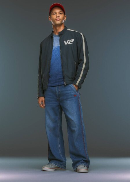 Uomo in piedi su sfondo sfumato, indossa giacca sportiva scura con logo “V2” sul petto, t-shirt blu sfumato, jeans larghi con banda laterale rossa, sneakers grigie e berretto rosso.<br /><br /><br /><br /><br /><br /><br /><br /><br /><br />
