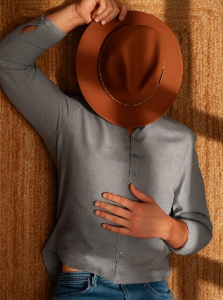 Persona sdraiata su un tappeto intrecciato color beige, con camicia grigia e jeans, mentre tiene il volto nascosto da un cappello marrone a tesa larga.