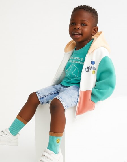 Bambino seduto su un blocco bianco in abbigliamento casual: felpa con cappuccio a blocchi di colore crema, verde menta e salmone, maglietta verde acqua con stampa, pantaloncini di jeans chiari, calzini verde tiffany e scarpe bianche, che sorride su sfondo neutro.<br /><br /><br /><br /><br /><br /><br /><br /><br /><br />