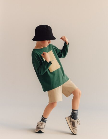 Bambino in piedi su sfondo neutro, con maglia verde a maniche lunghe, pantaloncini beige e cappello a secchiello nero, in posa dinamica con le braccia sollevate come in un gesto di energia.
