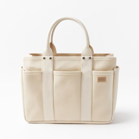 Borsa tote in tela beige chiaro con manici corti rinforzati e cuciture a vista. Il design è strutturato, con tasche esterne su tutti i lati e dettagli in tela tono su tono. Logo piccolo su etichetta marrone chiaro cucita frontalmente. Sfondo bianco uniforme.