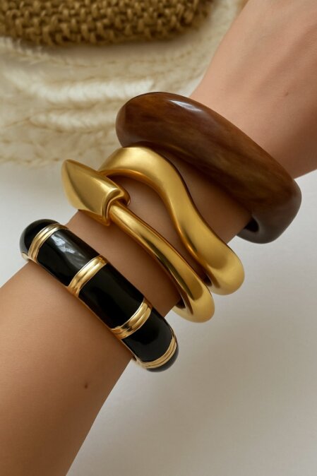Primo piano di un polso adornato con quattro bracciali rigidi di grandi dimensioni: uno in resina marrone scuro effetto legno, uno dorato con design ondulato, uno dorato con punta geometrica e uno nero lucido con inserti dorati in stile art déco. Sfondo neutro con texture intrecciata in alto.