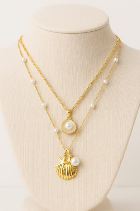 Collana dorata a doppio filo esposta su busto beige, composta da due catene sottili: la più corta presenta un pendente rotondo con perla centrale e dettagli martellati, mentre la più lunga ha ciondoli a forma di conchiglia e stella marina con perla laterale. Entrambe le catene sono decorate con perle distribuite a intervalli regolari.