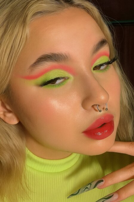 Primo piano di una giovane donna con trucco artistico e audace: ombretto verde lime brillante con dettagli sfumati rosa fluo allungati verso le tempie. Indossa rossetto lucido rosso, ha piercing al setto nasale e unghie lunghe decorate con motivo astratto verde. I capelli sono biondo chiaro e ondulati, e indossa un top a collo alto giallo fluorescente.<br /><br /><br /><br /><br /><br /><br /><br /><br /><br />
