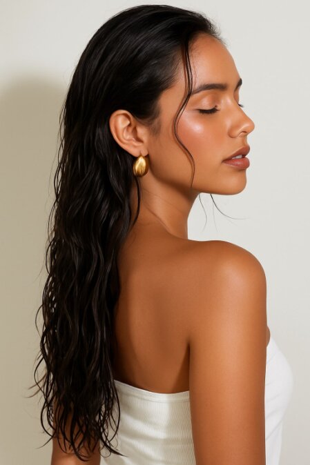 Donna con capelli lunghi e scuri dall’effetto bagnato, girata di profilo con occhi chiusi, indossa un top bianco senza spalline e un orecchino dorato. La pelle è luminosa e l’atmosfera elegante e raffinata.