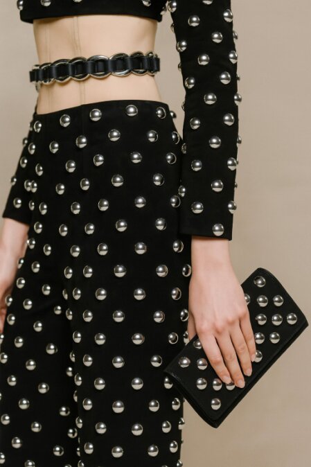 Modella con top corto e pantaloni neri decorati con borchie metalliche sferiche argentate, cintura abbinata e clutch nera con dettagli borchiati.