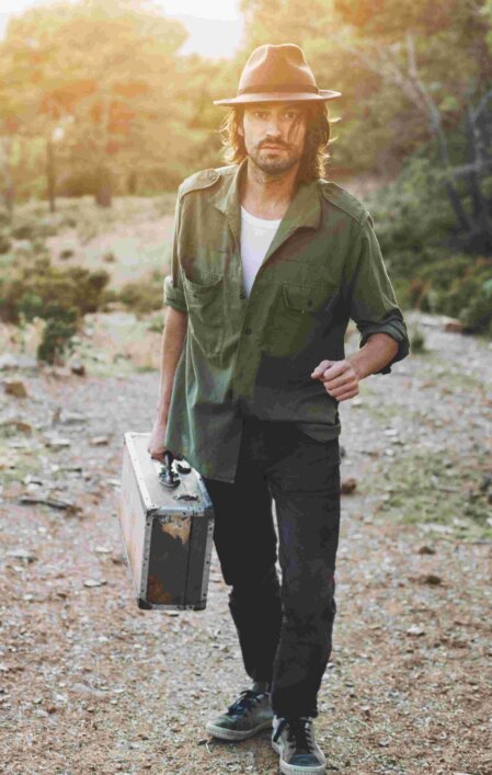 Uomo con barba e capelli lunghi che cammina in un sentiero sterrato tra la natura al tramonto, indossa una camicia verde militare con maniche arrotolate, pantaloni scuri, cappello marrone e porta una valigia vintage in metallo consumata.