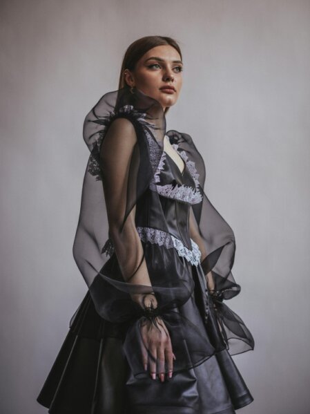 Donna con abito nero in ecopelle e dettagli in tulle trasparente e pizzo bianco, in posa elegante su sfondo neutro in studio.