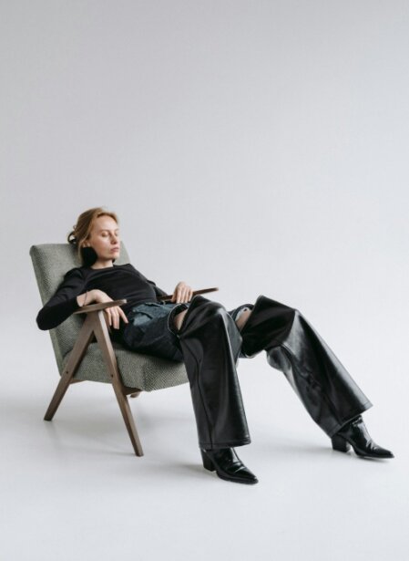 Donna bionda con outfit nero e pantaloni in pelle, seduta in modo rilassato su una poltrona di design in uno studio fotografico minimalista con sfondo bianco.