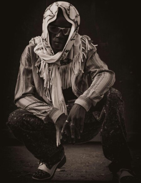 Uomo accovacciato in posa, indossa occhiali da sole, foulard bianco con disegni neri e camicia a righe, in uno scatto in bianco e nero con forte contrasto.