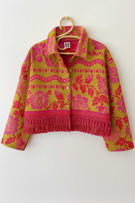 Giacca corta appesa a una gruccia rotonda, realizzata in tessuto jacquard giallo senape con motivi floreali rosa e rossi. Il capo presenta una chiusura a bottoni, colletto classico e una vistosa frangia rossa decorativa lungo l’orlo inferiore. Il design è vivace e ricco di dettagli tessili.<br /><br /><br /><br /><br /><br /><br /><br /><br /><br />Chiedi a ChatGPT<br />