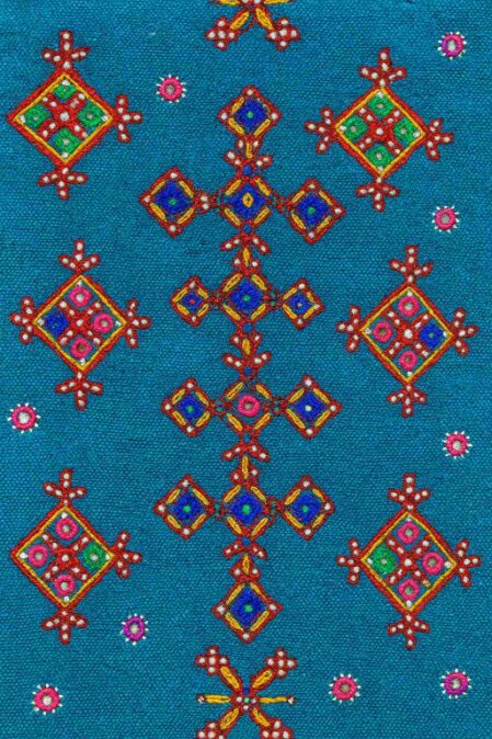 Dettaglio di un tessuto azzurro decorato con un ricamo geometrico e floreale multicolore. Il motivo centrale è formato da una serie di rombi blu con bordi rossi e gialli, disposti in croce, mentre i motivi laterali includono rombi più grandi con motivi interni verdi, rosa e bianchi. Piccoli cerchi floreali rosa e viola punteggiano lo sfondo, creando un effetto ornamentale complesso e vibrante.