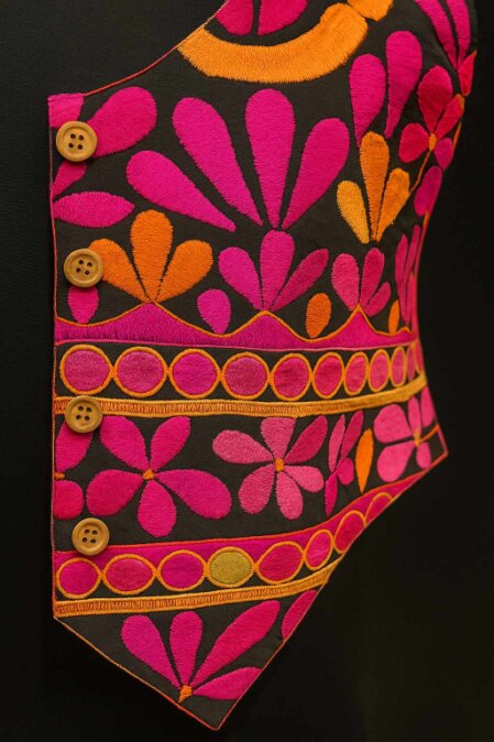 Primo piano di un gilet decorato con ricami floreali dai toni vivaci del fucsia, arancione e rosa su fondo nero. Il capo è impreziosito da quattro bottoni in legno sul lato sinistro e presenta motivi stilizzati a forma di fiori e cerchi lungo l’orlo inferiore, creando un effetto visivo dinamico e colorato.