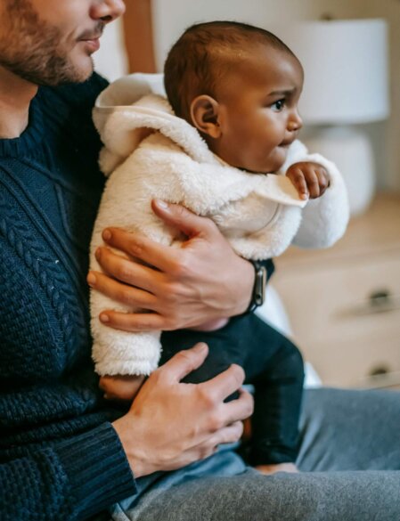 Primo piano di un uomo con maglione blu scuro e pantaloni grigi che tiene in braccio un bambino con giacca sherpa bianca e pantaloni neri, seduti in un ambiente domestico con arredi chiari.