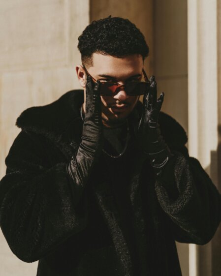 Uomo elegante con cappotto nero in pelliccia, guanti in pelle e occhiali da sole, in posa all’aperto vicino a un edificio in pietra, illuminato da luce naturale.
