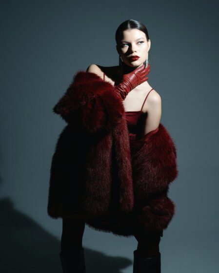 Modella con cappotto di pelliccia rosso scuro e guanti in pelle rossa, posa con eleganza su sfondo grigio scuro, illuminata da luce direzionale che crea un'atmosfera drammatica.