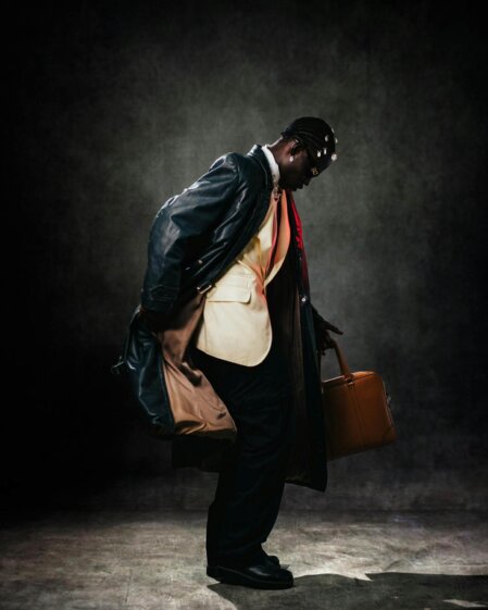 Uomo in posa con cappotto lungo nero, blazer color crema e cravatta rossa, mentre tiene una borsa marrone in mano su sfondo scuro.