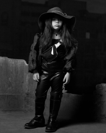 Bambina in abiti neri e stivali alti, con cappello a tesa larga e marsupio a tracolla, fotografata in bianco e nero su uno sfondo urbano notturno.
