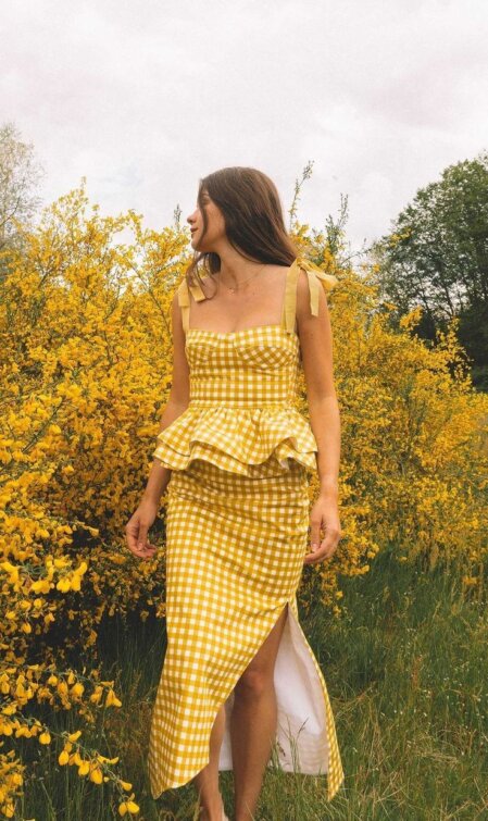 Donna vista a mezzo busto e gambe in un abito lungo a quadretti gialli con spalline annodate e balza in vita, in piedi davanti a cespugli di fiori gialli in fiore.