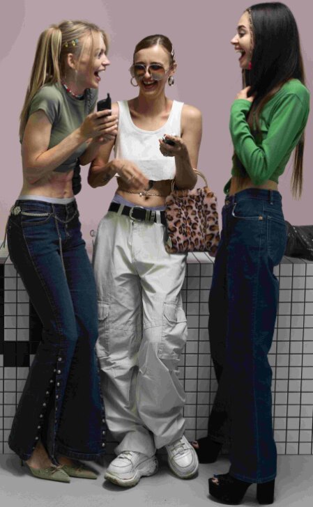 Tre ragazze vestite in stile anni 2000 ridono insieme in un ambiente urbano; indossano crop top, pantaloni a vita bassa, accessori appariscenti come cinture con catene e borsa leopardata, e acconciature con clip colorate, incarnando lo spirito nostalgico e spensierato della moda Y2K.