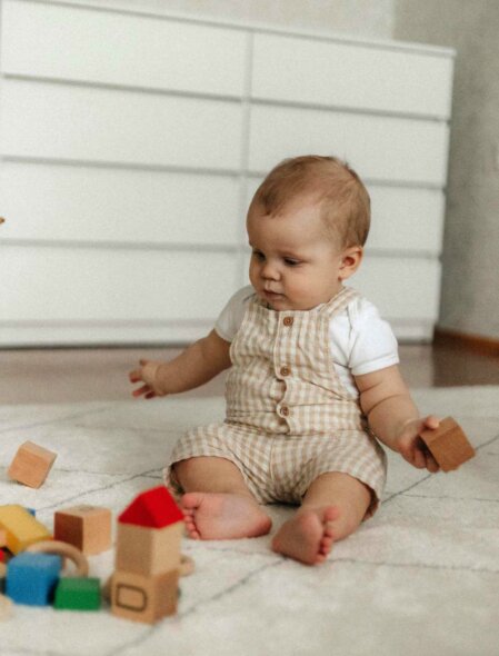 Neonato seduto su un tappeto beige che gioca con cubi di legno colorati, indossa una salopette a quadretti beige e bianca con bottoni, in un ambiente minimalista con cassettiera sullo sfondo, stile moda neonato naturale e giocoso.