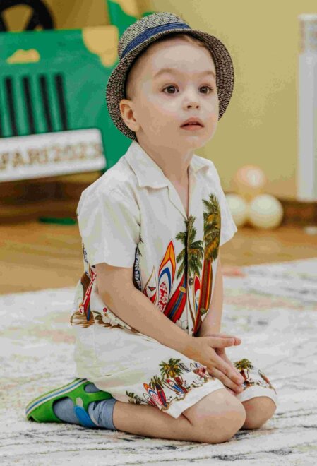 Bambino seduto sul tappeto con espressione incuriosita, indossa un completo estivo con stampa di palme e tavole da surf, cappello di paglia a tesa corta e scarpe verdi con calzini azzurri, in un ambiente interno colorato e giocoso.
