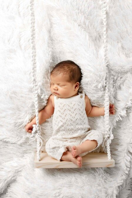 Neonato che dorme su altalena in legno, indossa tutina in maglia beige con motivo traforato, ambientazione soffice su coperta bianca, ideale per campagna moda neonato estate.