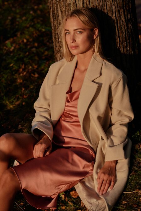 Donna seduta sull’erba accanto a un albero, indossa un abito in raso color rosa cipria e un cappotto avorio. La scena è illuminata dalla luce calda del tramonto, che crea contrasti morbidi sul viso e sull’outfit.