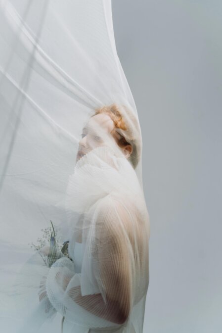Donna con capelli rossi avvolta in un velo trasparente bianco, che copre il suo viso e l'abito elegante con colletto plissettato. Tiene in mano un piccolo bouquet floreale, immersa in una luce soffusa su sfondo grigio chiaro.