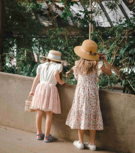 Due bambine di spalle con abiti estivi floreali e cappelli di paglia osservano la vegetazione in una serra, una con borsetta intrecciata e l’altra con sneaker bianche, moda bambina primavera-estate romantica e naturale.