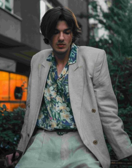 Giovane uomo seduto all’aperto con espressione riflessiva, indossa una camicia hawaiana a fiori dai toni verdi e crema abbinata a un blazer beige con spalle strutturate e pantaloni chiari, in un contesto urbano con vegetazione e luci calde sullo sfondo.