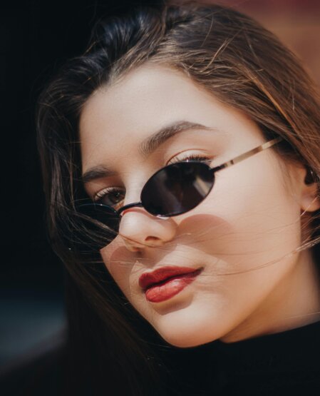 Ritratto ravvicinato di una donna con lunghi capelli castani, indossa occhiali da sole ovali scuri e un rossetto rosso intenso. Lo sguardo è diretto verso l’obiettivo e il viso è parzialmente illuminato dalla luce solare, che crea ombre morbide. Indossa un dolcevita nero.