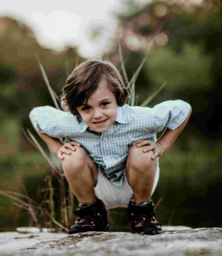 Bambino con capelli castani e camicia a quadretti azzurri accovacciato su una roccia all’aperto, con pantaloncini chiari e scarpe da trekking nere e rosse, immerso in un contesto naturale sfocato sullo sfondo.