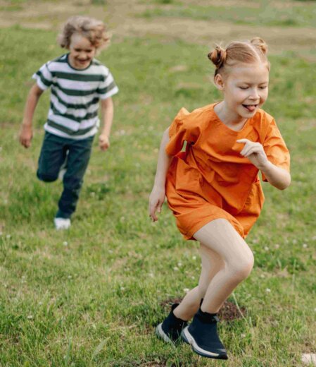 Bambina con vestito arancione acceso e capelli raccolti in due chignon che corre sorridente su un prato, seguita da un bambino con maglietta a righe bianche e verdi e pantaloni scuri, durante un gioco all’aperto.