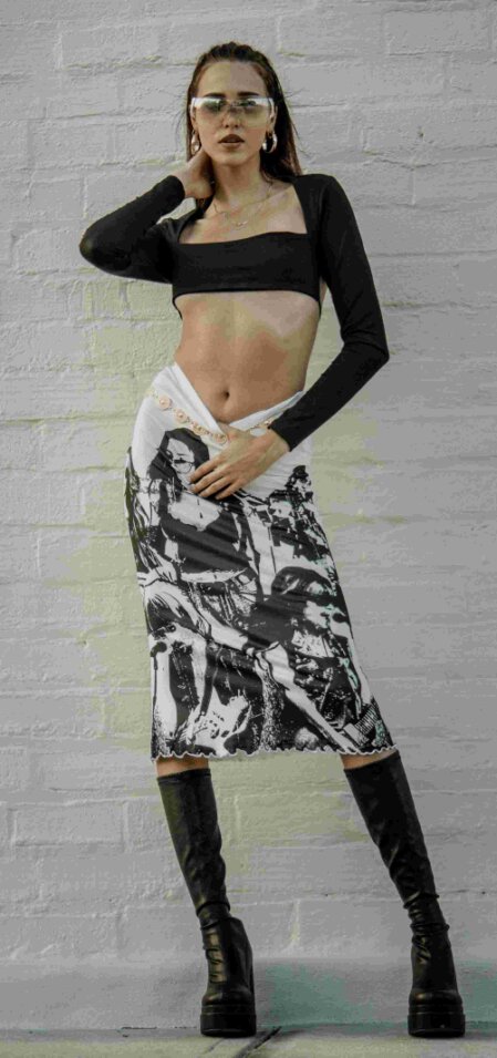 Modella in posa contro un muro bianco, indossa un crop top nero a maniche lunghe, una gonna bianca e nera con stampa grafica artistica e stivali alti in pelle nera; occhiali da sole trasparenti e orecchini a cerchio completano il look moderno e audace.