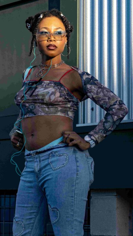 Giovane donna posa con sicurezza indossando un crop top a maniche lunghe con fantasia floreale e pantaloni di jeans a vita bassa decorati con cuori cuciti; completa il look con accessori audaci come occhiali da sole rettangolari, ombretto azzurro acceso, orecchini a cerchio e collane a catena, richiamando lo stile streetwear anni 2000.