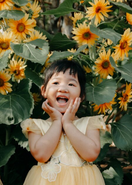 Bambina sorridente con le mani appoggiate alle guance, in un abito giallo decorato, davanti a un folto gruppo di girasoli in piena fioritura.
