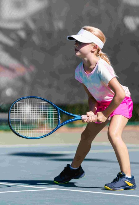 Bambina bionda con coda di cavallo e visiera bianca pronta a rispondere in una partita di tennis, impugnando una racchetta blu sul campo all’aperto, con outfit sportivo rosa e scarpe da ginnastica nere.<br /><br /><br /><br /><br /><br /><br /><br /><br /><br />