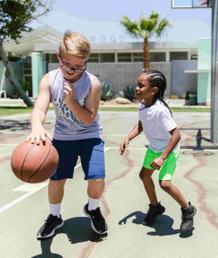 Due bambini giocano a basket all’aperto su un campo soleggiato; il bambino con capelli biondi, occhiali e maglietta grigia palleggia il pallone mentre l’altro bambino con treccine e pantaloncini verde fluo tenta di difendere.
