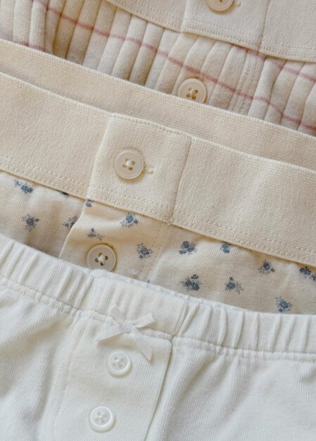 Capi intimi impilati in toni neutri con vita elasticizzata: la prima in tessuto coste beige con sottili righe rosa e bottone frontale, la seconda panna con piccoli motivi floreali azzurri e bottone centrale, la terza bianca con fiocchetto decorativo e bottoni frontali, visibili dettagli di cuciture ed elastico in vita.