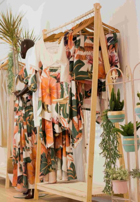 Espositore in legno con abiti coordinati a fantasia floreale tropicale nei toni dell’arancione, rosa e verde, esposti su grucce accanto a un manichino vestito in stile simile; sullo sfondo piante decorative in vasi sospesi contribuiscono a creare un’atmosfera naturale e accogliente all’interno di una boutique.