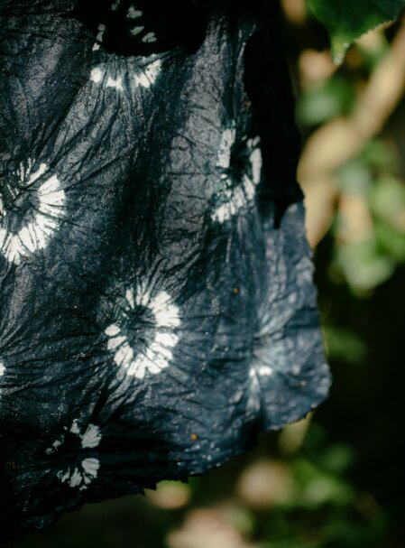 Dettaglio ravvicinato di un tessuto nero con motivo tie-dye bianco, caratterizzato da cerchi concentrici irregolari. La stoffa appare leggermente stropicciata e viene illuminata da luce naturale, con un fondale sfocato di foglie verdi e rami.