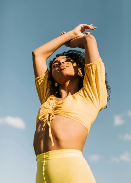 Persona adulta con capelli ricci scuri, in piedi all’aperto sotto un cielo sereno, indossa un top giallo corto con coulisse centrale e leggings gialli, con le braccia sollevate sopra la testa e un piccolo fiore giallo vicino al viso, espressione rivolta verso il sole.