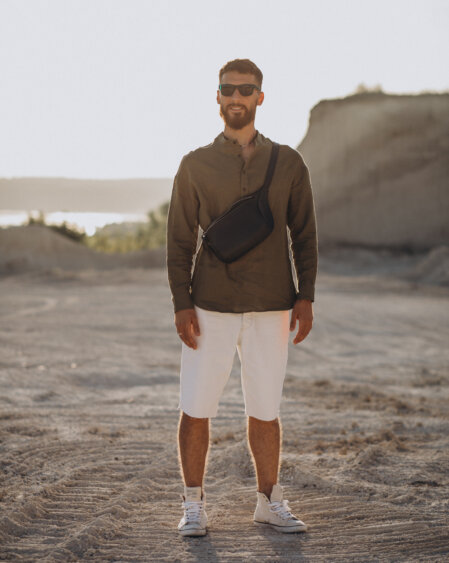 Uomo in piedi su un terreno sabbioso sotto la luce del tramonto, indossa una camicia verde oliva a maniche lunghe, pantaloncini bianchi al ginocchio, scarpe sportive bianche e una tracolla nera. Indossa occhiali da sole e sorride leggermente. Sullo sfondo, un paesaggio roccioso e naturale.